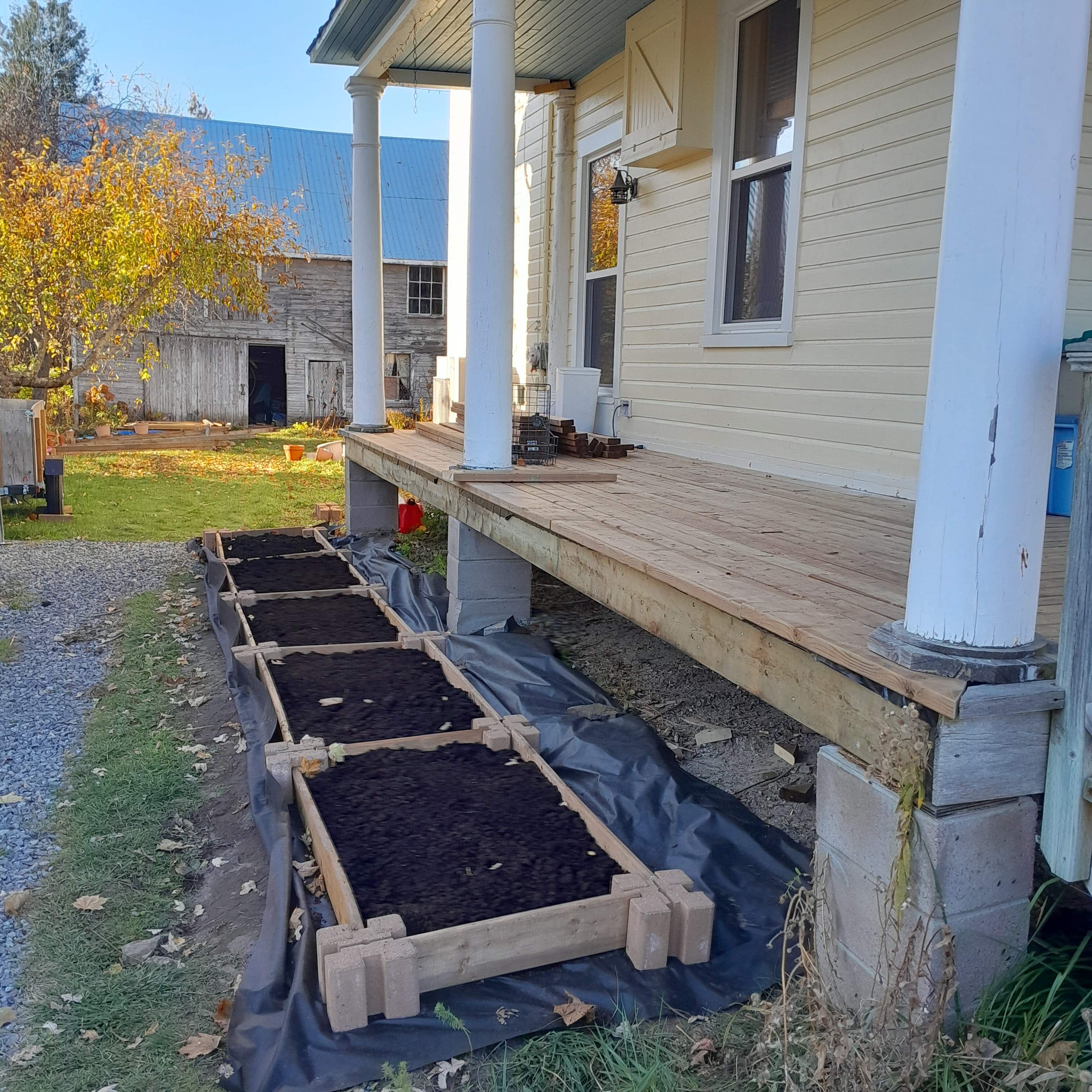 The image depicts a house with a garden bed in front of it. The house is yellow with white columns and has a wooden porch....